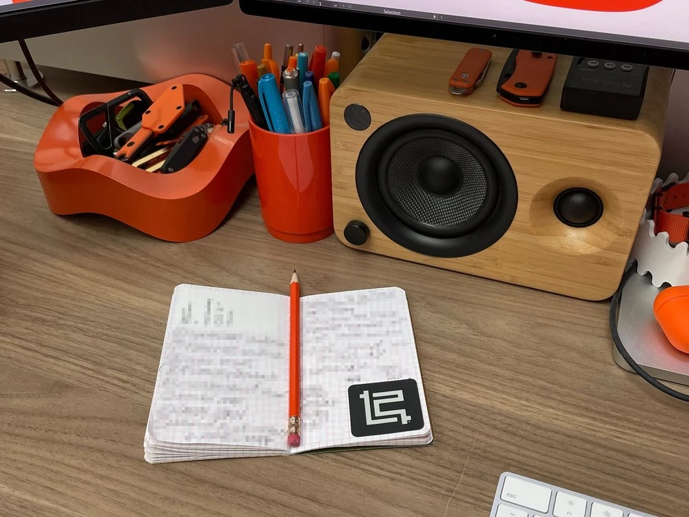 Aaron Draplin's desktop with orange Helit Sinus organizers, open Field Notes notebook, pencil, and Kanto wooden speaker