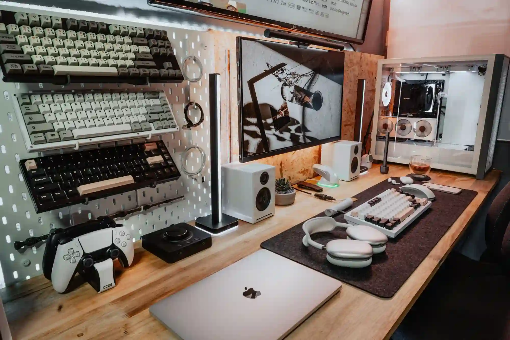 TheBucketListTech's desk setup with multiple keyboards mounted on a pegboard, custom PC build, LG monitor, MacBook, speakers, and PS5 controller